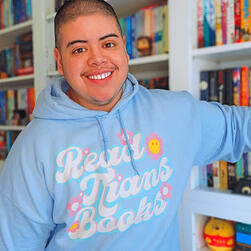 A photo of Adri—a Mexican-American transmasc nonbinary person with a star-shaped barbell in their right eyebrow—standing in front of their white bookshelves. They’re wearing a powder blue hoodie that says “read trans books” in big white, looping letters ac