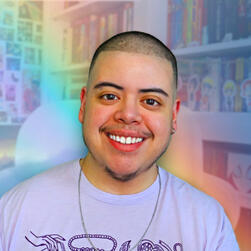 A photograph of Adri smiling and sitting in front of their bookshelves. They're wearing a light purple t-shirt, a silver chain necklace, a silver hoop earring in their left ear, and a silver barbell with a cluster of stars in their right eyebrow. The backg